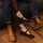 Pair of black suede Lola Mary Jane flats with gold buckles worn with denim jeans and brown leather boots.